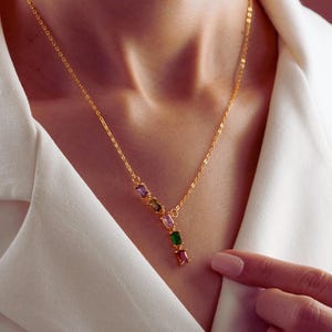 May include: A gold-tone chain necklace featuring a vertical drop pendant composed of five small, rectangular, multi-colored gemstones in shades of green, pink, and purple, resting against a white collared garment.