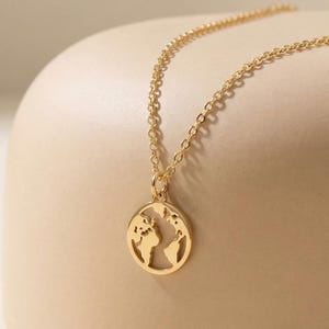 May include: A gold necklace with a circular pendant depicting a world map. The pendant is suspended from a fine chain. The jewellery is set against a neutral backdrop, emphasizing its simple elegance.