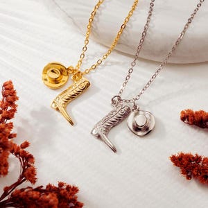 Könnte beinhalten: Zwei Halsketten mit Anhängern im Cowboy-Stil. Eine ist goldfarben mit einem Cowboy-Stiefel und einem Hut-Charm. Die andere ist silberfarben mit einem Cowboy-Stiefel und einem Hut-Charm. Die Halsketten liegen auf einer weißen Oberfläche mit getrockneten Blumen.