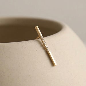 May include: A gold bar earring with a line of small, clear gemstones. The earring is a simple, modern design, and is displayed against a neutral background.