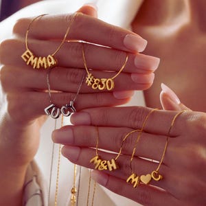Könnte beinhalten: Goldfarbene Halsketten mit Buchstaben- und Zahlenanhängern, die "EMMA", "#830", "LUCY", "M&H" und "M♡O" buchstabieren. Die Halsketten werden vor einem weichen Hintergrund präsentiert, der den personalisierten Schmuck hervorhebt.