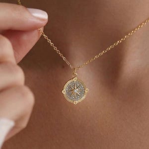May include: A gold necklace featuring a compass pendant. The pendant is circular with a starburst design, incorporating a central clear stone and smaller surrounding stones. The delicate chain is worn against a skin tone background.