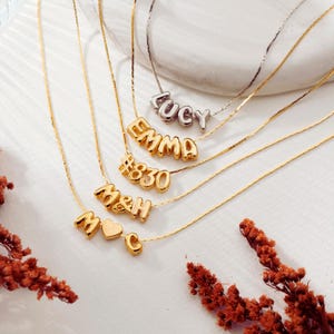 May include: Five delicate necklaces with thin gold and silver chains. The pendants feature metallic bubble-style letters and numbers spelling Lucy, Emma, #830, M&H, and M heart C. Each piece is crafted with a polished finish and a simple clasp.