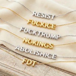 May include: A collection of six delicate necklaces featuring silver and gold-toned metal chains. Each necklace displays a central word or acronym pendant in block letters: RESIST, FUCKICE, FUCKTRUMP, NOKINGS, ABOLISHICE, and FDT, crafted in matching metal.