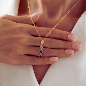 May include: A gold-tone chain necklace featuring a vertical pendant with four small heart-shaped gemstones in clear, light pink, blue, and purple colors, arranged in a descending line on a delicate link chain.