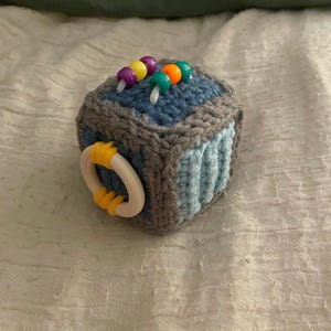 May include: A handmade crochet cube toy in shades of blue, gray, and white. The cube features colorful beads and a white ring with yellow accents. The toy is designed for sensory play and is likely for babies or toddlers.