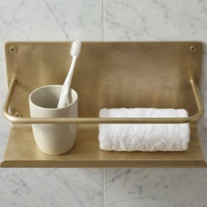 Könnte beinhalten: Messing-Badezimmerregal mit Zahnbürste in einem Keramikbecher und einem gerollten weißen Handtuch. Das Regal hat eine gebogene Stange und ist an einer Marmorfliesenwand befestigt. Modernes Design.
