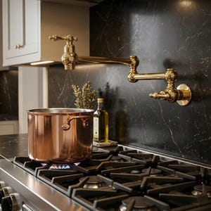 May include: A copper pot sits on a gas stove with a blue flame. A gold-colored faucet is mounted on the black marble backsplash. The kitchen setting includes a cabinet and olive oil bottles.