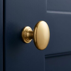 K&ouml;nnte beinhalten: Ein goldfarbener, ovaler Schrankknauf mit kreisf&ouml;rmiger Basis, montiert an einer dunkelblauen Schrankt&uuml;r. Der Knauf hat eine glatte, polierte Oberfl&auml;che. Diese Hardware ist f&uuml;r K&uuml;chen- oder Badezimmerschr&auml;nke geeignet. Ma&szlig;e in Zentimetern.