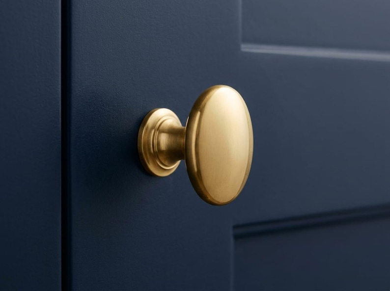 K&ouml;nnte beinhalten: Ein goldfarbener, ovaler Schrankknauf mit kreisf&ouml;rmiger Basis, montiert an einer dunkelblauen Schrankt&uuml;r. Der Knauf hat eine glatte, polierte Oberfl&auml;che. Diese Hardware ist f&uuml;r K&uuml;chen- oder Badezimmerschr&auml;nke geeignet. Ma&szlig;e in Zentimetern.