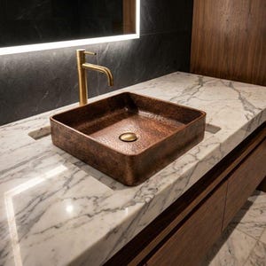 May include: A square, hammered copper sink with a brass drain and faucet. The sink is set on a white marble countertop with a dark wood cabinet below. A backlit mirror is above the sink, and the faucet is gold-colored.