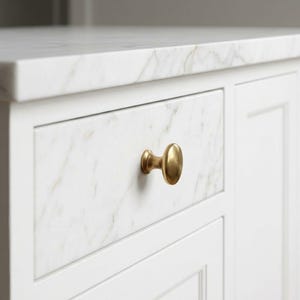 K&ouml;nnte beinhalten: Nahaufnahme einer wei&szlig;en K&uuml;cheninsel mit Marmor-Arbeitsplatte. Die Schublade hat eine Marmorfront und einen goldfarbenen Knauf. Die Insel hat ein klares, modernes Design.