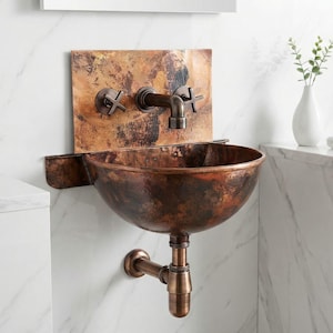 Puede incluir: Un lavabo de baño de color cobre con un grifo y una tubería de desagüe a juego. El lavabo tiene forma de cuenco y está montado en una pared con diseño de mármol. El grifo tiene manijas en forma de cruz. Un jarrón blanco con vegetación se encuentra sobre una encimera de mármol.