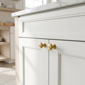 K&ouml;nnte beinhalten: Nahaufnahme eines wei&szlig;en K&uuml;chenschranks mit zwei goldfarbenen runden Kn&ouml;pfen. Die Schrankt&uuml;ren haben ein eingelassenes Paneeldesign. Im Hintergrund sind ein helles Holzregal und eine Arbeitsplatte zu sehen.