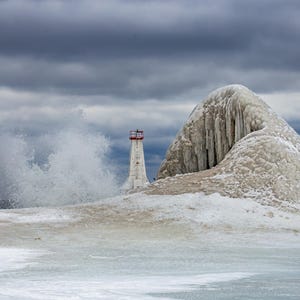 Op de afbeelding: Een witte vuurtoren met een rode top staat tegen een stormachtige lucht, met golven die in de buurt breken. De vuurtoren is omgeven door ijs en sneeuw, met een grote ijsformatie aan de rechterkant. De scène roept een koude, kustachtige omgeving op.