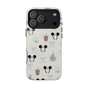 May include: A phone case with a cream background featuring a repeating pattern of black bow-shaped mouse ears, blue bows, coffee drinks, genie lamps, and castle illustrations. The case has a black camera module.