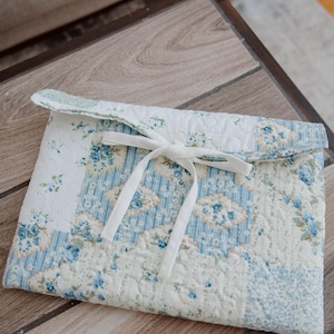 May include: A quilted fabric sleeve with a floral pattern in shades of blue and white. The sleeve is secured with a white ribbon tie. The design features a patchwork style with various floral motifs.