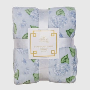 May include: A folded, light blue throw blanket with a floral pattern of blue hydrangeas and green leaves. The blanket is tied with a white ribbon and has a tag that reads "Luxuriously Soft Throw" and measures 50" x 60".
