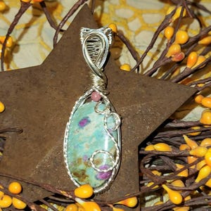 May include: A handcrafted pendant featuring a turquoise-colored stone with red and brown inclusions, wrapped in silver-colored wire. A small pink bead accents the top. The pendant is suspended from a silver-colored wire loop.