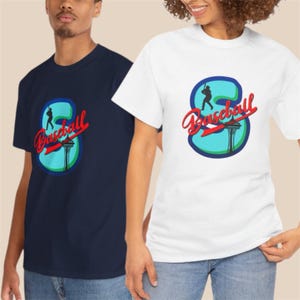 Pode incluir: Duas camisetas, uma azul marinho e outra branca, cada uma com um design com tema de beisebol. O design inclui a palavra "Baseball" em script vermelho, uma silhueta de um batedor e o Space Needle. As camisetas são de gola redonda.