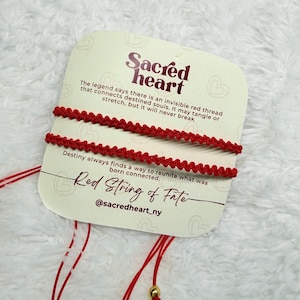 Könnte beinhalten: Zwei rote Schnurarmbänder werden auf einer weißen Karte gezeigt. Auf der Karte steht "Sacred Heart" und "Red String of Fate", sowie Text über das Schicksal. Die Armbänder sind geknotet.