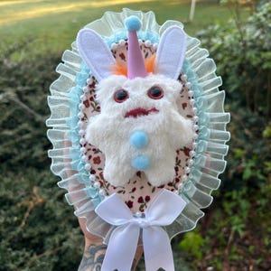 May include: A handmade plush creature with white fur, large red eyes, and a party hat. The creature is set on an oval-shaped base with a floral pattern, surrounded by blue lace and a white bow.