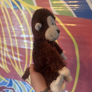 Könnte beinhalten: Ein brauner Plüschaffe mit hellbraunem Gesicht, Ohren, Händen und Füßen. Das Stofftier wird in einer Hand gehalten und hat einen kleinen Schwanz. Der Affe ist ein weiches Spielzeug.