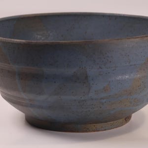 May include: A handmade ceramic bowl with a speckled blue glaze. The bowl has a wide, rounded shape with a textured exterior and a footed base. The rim and base have a darker, earthy tone.