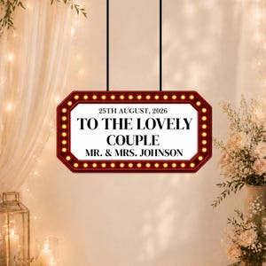 May include: A rectangular sign with a red border and small lights, hanging from black cords. The sign reads "25th August, 2026" and "TO THE LOVELY COUPLE MR. & MRS. JOHNSON" in a classic font. The background is a soft, warm color.