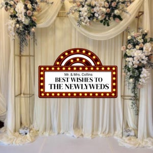 May include: A wedding backdrop featuring a sign that reads "Mr. & Mrs. Collins BEST WISHES TO THE NEWLYWEDS." The sign is framed with a marquee-style border of lights. The backdrop includes white draping, floral arrangements, and gold metal structures.