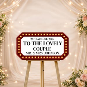May include: A red and gold sign on a wooden easel with the text "TO THE LOVELY COUPLE MR. & MRS. JOHNSON" and the date "25TH AUGUST, 2026". The sign is surrounded by small lights, with a floral and curtain backdrop.