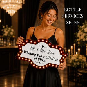 May include: A decorative sign with a red border and illuminated bulbs. The sign has a white center with the text "Mr. & Mrs. Steve Wishing You a Lifetime of Bliss." The sign is held by a person in a black dress.