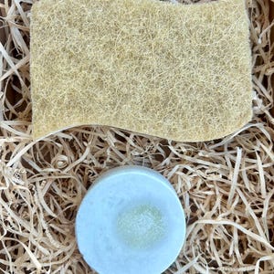 Può includere: Una spugna di luffa beige rettangolare dai bordi ondulati posizionata sopra una saponetta circolare azzurro pallido con un centro testurizzato verde chiaro, il tutto su un letto di sottili trucioli di legno marrone chiaro per imballaggio.