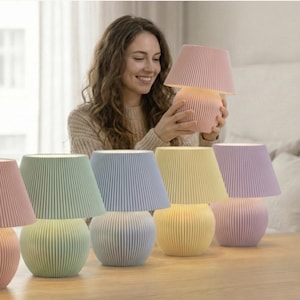 May include: A selection of pastel-coloured table lamps with pleated shades and rounded bases. The lamps are in shades of pink, green, blue, yellow, and purple. The lamps are arranged on a wooden table.