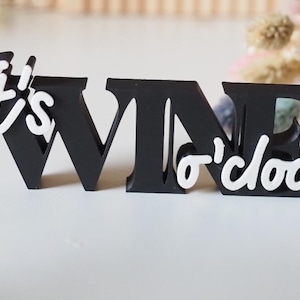 May include: Black wooden sign with the words "It's Wine o'clock" in a playful font. The words "It's" and "o'clock" are in white, contrasting with the black letters of "WINE". The sign is a decorative item.