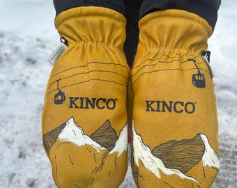 May include: Mustard yellow mittens with the brand name "KINCO" printed in black. The mittens feature a mountain range graphic with white snow-capped peaks and a ski lift design. The mittens are worn over black pants.