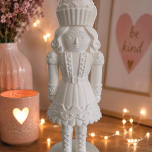 May include: A matte white nutcracker figurine featuring a cupcake-shaped hat with a cherry on top. The figure wears a uniform with a belted waist, a skirt with heart-shaped details, and tall boots, all standing on a circular base with decorative trim.
