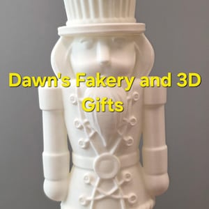 May include: A white 3D-printed figurine of a nutcracker soldier with a cupcake-shaped hat. The figurine has detailed features, including buttons, a belt, and boots. The text "Dawn's Fakery and 3D Gifts" is visible.