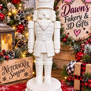 May include: A solid white nutcracker figurine standing on a circular base. The nutcracker features a tall hat, a detailed uniform with epaulets, and a beard. The design includes intricate embossed patterns on the jacket, sleeves, and boots.