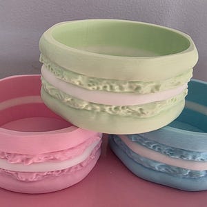 Puede incluir: Tres recipientes decorativos de colores pastel con forma de macarons. Los recipientes son en tonos rosa, verde y azul, con una capa blanca en el medio. Los recipientes están sobre una superficie rosa.