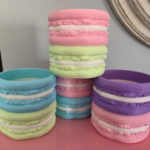 Puede incluir: Una colección de maceteros de gran tamaño con forma de macaron y colores pastel. Los maceteros son de color rosa, azul, verde y morado, con detalles blancos. Estos maceteros decorativos añaden un toque caprichoso a cualquier espacio.