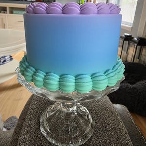 Puede incluir: Un pastel redondo con glaseado degradado azul y morado, decorado con glaseado en la parte superior e inferior. El pastel se encuentra sobre un soporte de cristal transparente con borde festoneado.
