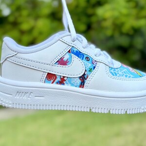 K&ouml;nnte beinhalten: Wei&szlig;er Nike Air Force 1 Sneaker mit farbenfrohem Spider-Man-Design auf den Seitenteilen. Der Schuh hat eine wei&szlig;e Sohle, Schn&uuml;rsenkel und das Nike Swoosh-Logo. Das Wort "NIKE" ist auf der Seite der Sohle aufgedruckt.