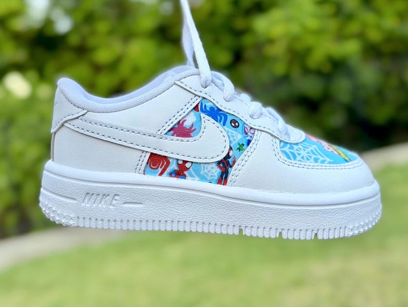 K&ouml;nnte beinhalten: Wei&szlig;er Nike Air Force 1 Sneaker mit farbenfrohem Spider-Man-Design auf den Seitenteilen. Der Schuh hat eine wei&szlig;e Sohle, Schn&uuml;rsenkel und das Nike Swoosh-Logo. Das Wort "NIKE" ist auf der Seite der Sohle aufgedruckt.