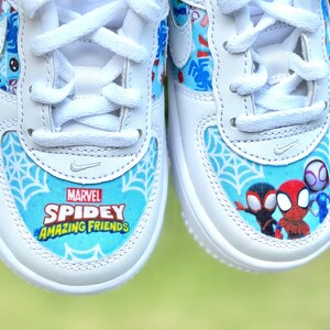 K&ouml;nnte beinhalten: Wei&szlig;e Turnschuhe mit blau-wei&szlig;en Spidey-Grafiken. Die Zehenpartie zeigt den Text "Marvel Spidey Amazing Friends" &uuml;ber einem Spinnennetz-Design. Die Seiten haben Cartoon-Bilder von Spidey-Charakteren.