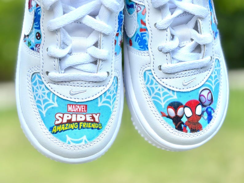 K&ouml;nnte beinhalten: Wei&szlig;e Turnschuhe mit blau-wei&szlig;en Spidey-Grafiken. Die Zehenpartie zeigt den Text "Marvel Spidey Amazing Friends" &uuml;ber einem Spinnennetz-Design. Die Seiten haben Cartoon-Bilder von Spidey-Charakteren.