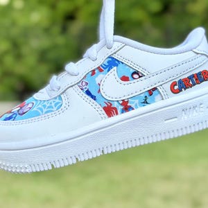 K&ouml;nnte beinhalten: Wei&szlig;er Nike Air Force 1 Sneaker mit individuellem Spider-Man-Design. Der Schuh hat ein blaues und rotes Muster mit Spider-Man-Grafiken. Der Name "CARTIER" ist rot und blau auf der Seite aufgedruckt.