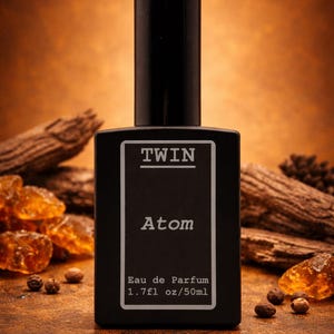 May include: A black rectangular perfume bottle with a black cap. The label reads "TWIN Atom Eau de Parfum 1.7 fl oz/50ml". The background is a warm brown with wood and amber accents.