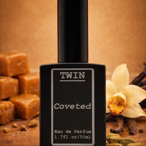 May include: A black perfume bottle with a rectangular shape and a tall, cylindrical cap. The bottle is labeled "TWIN" and "Coveted" with "Eau de Parfum 1.7fl oz/50ml". The background includes vanilla, cardamom, and caramel candies.