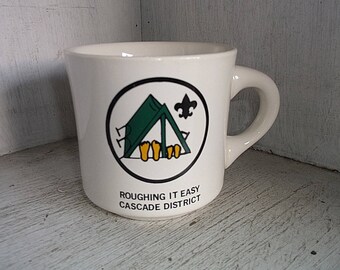Puede incluir: Taza de cerámica blanca con un logotipo verde y negro que representa una tienda de campaña y el texto "Roughing It Easy Cascade District".